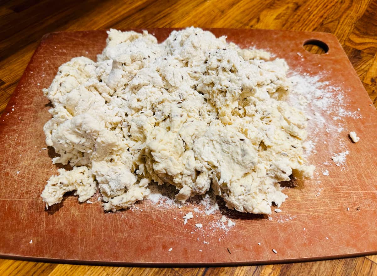 Rough clumps of pale yellow dough flecked with caraway seeds on a cutting board.