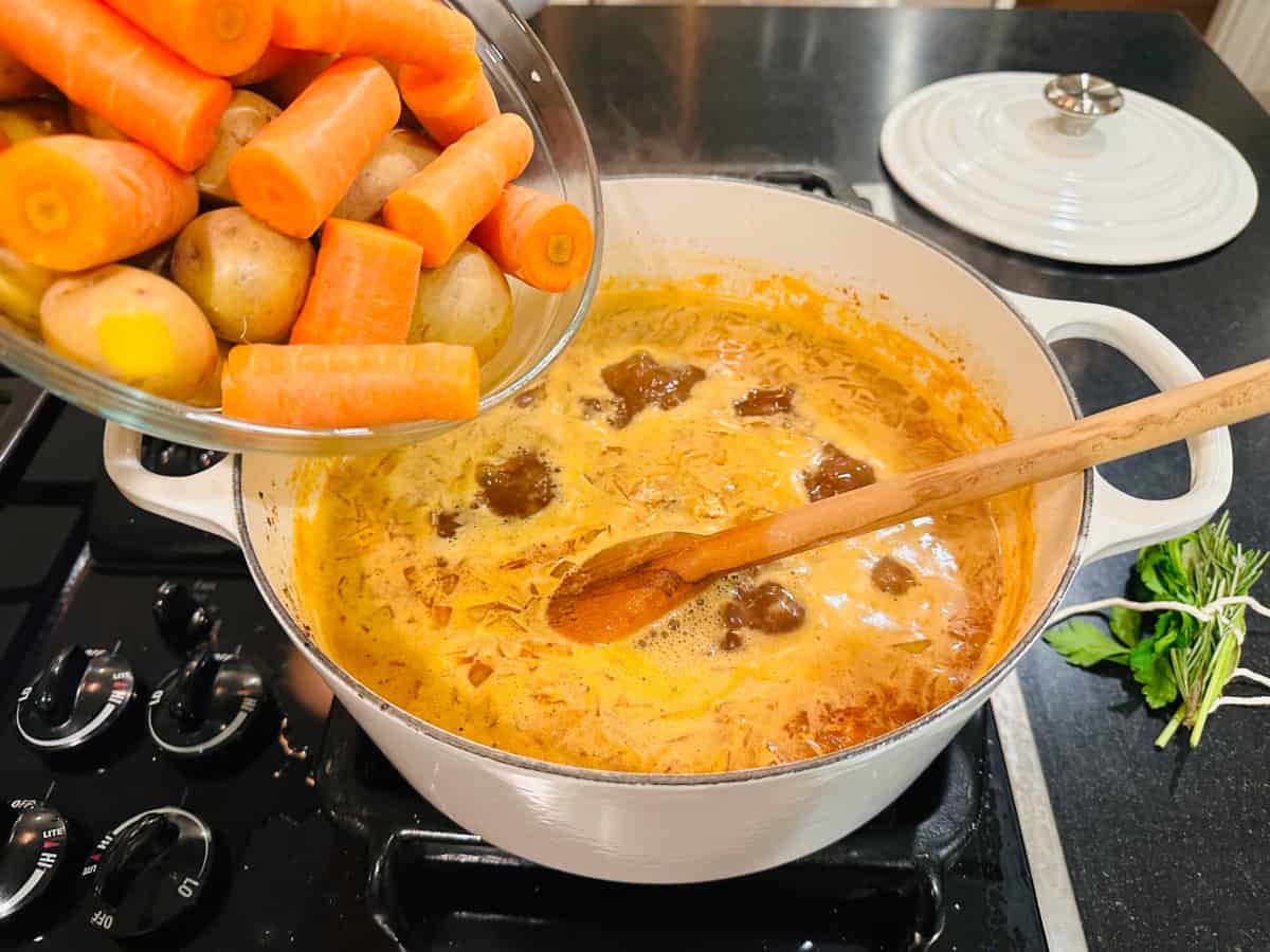Brown liquid in a white pot with a wooden spoon along with a glass bowl of carrots and potatoes.