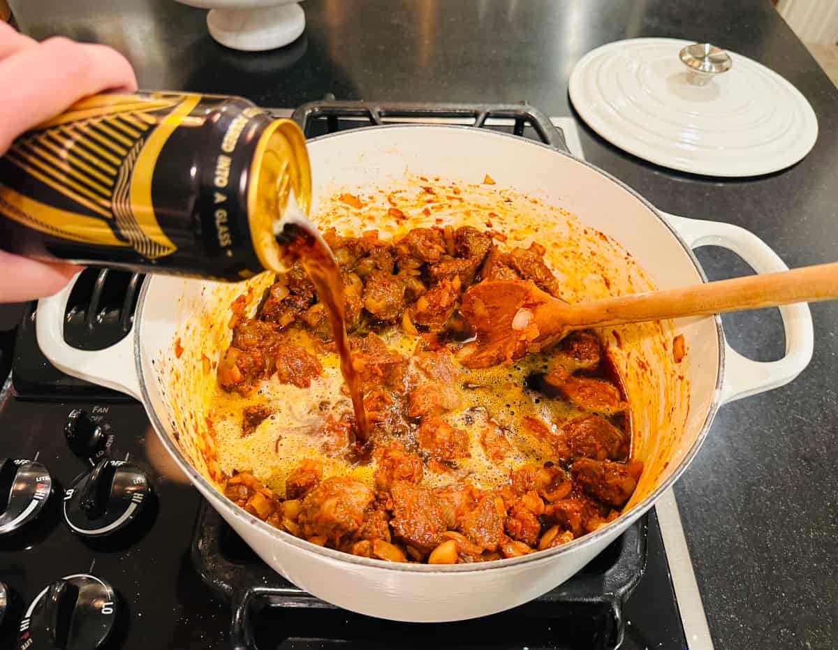 Can of Guinness stout being poured over browned lamb pieces in a white pot with a wooden spoon.