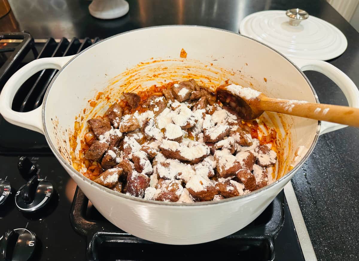 Browned lamb sprinkled with flour in a white pot with a wooden spoon.