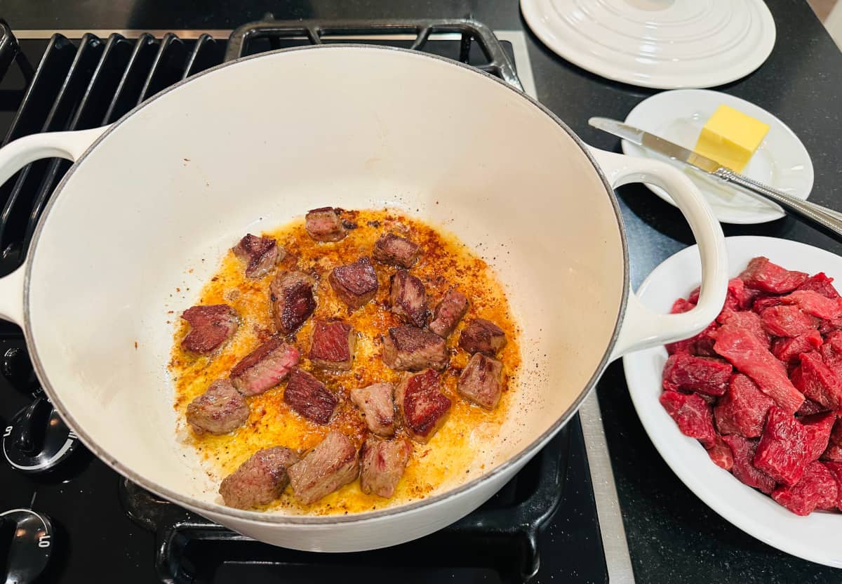 Lamb pieces browning in a white pot next to a white plate of raw lamb pieces and a small plate of butter.