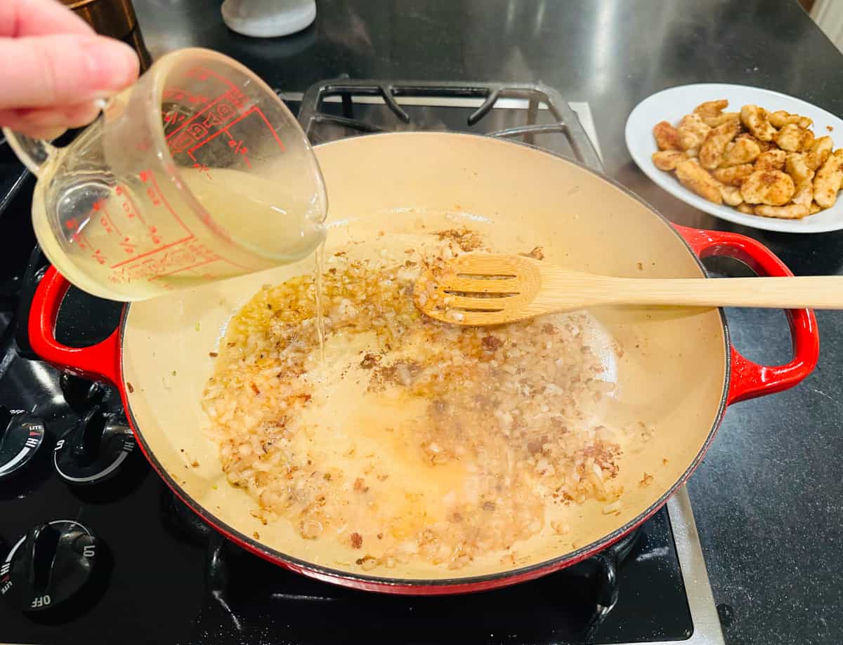 Wine being poured from a measuring cup into a wide pan containing browned shallots and a wooden spoon.