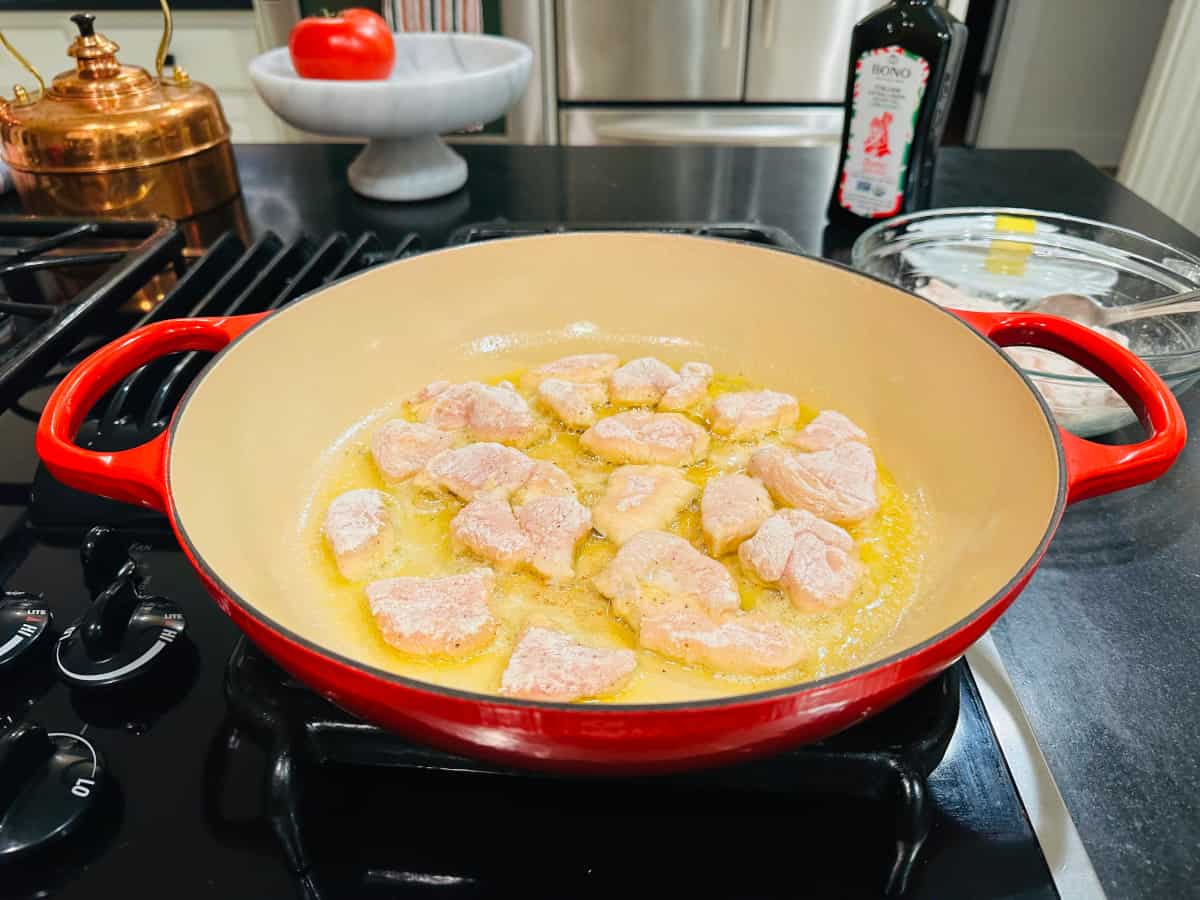Small pieces of chicken frying in a wide pan with red handles.
