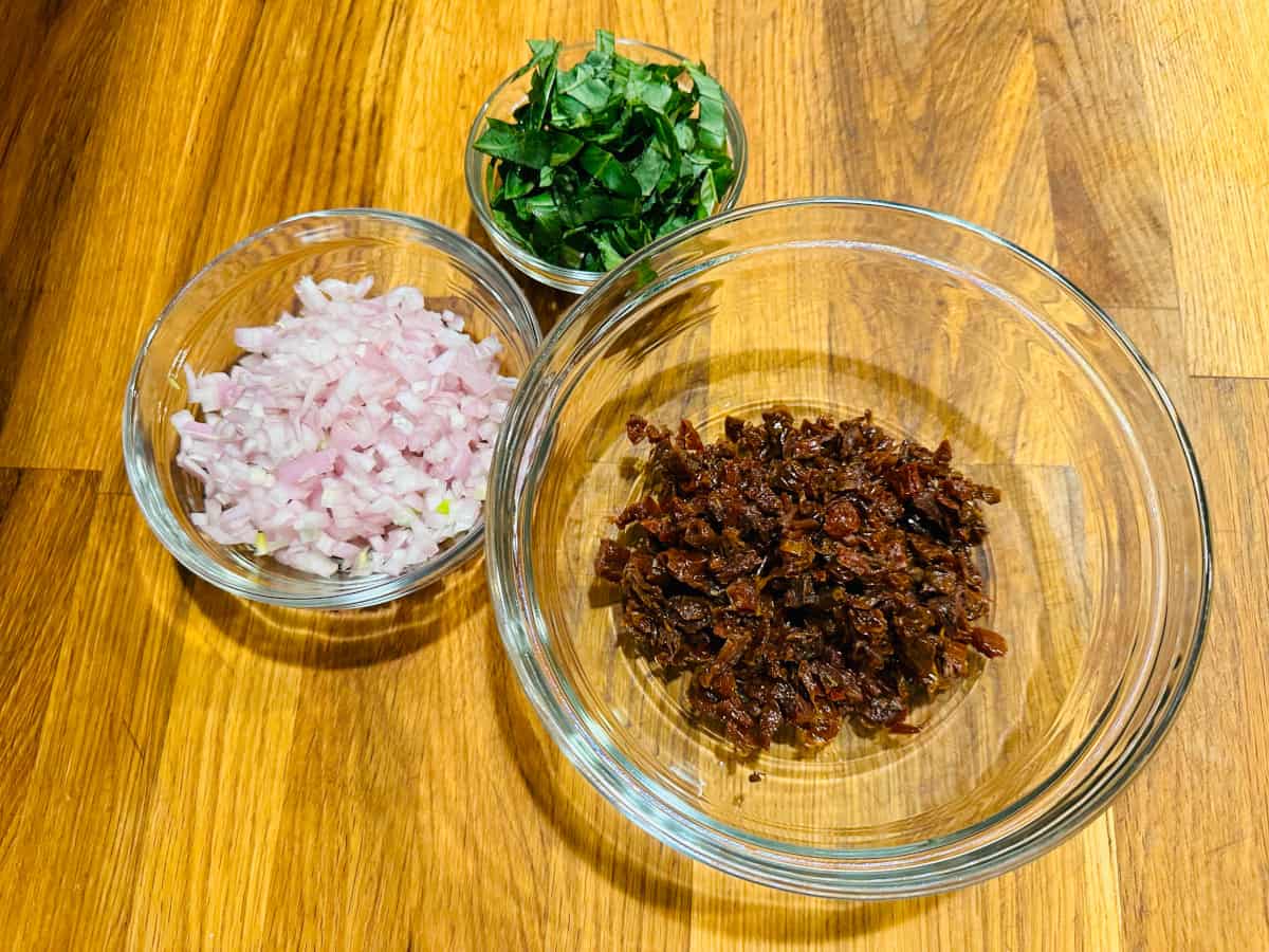 Three separate glass bowls containing chopped shallots, basil, and sun-dried tomatoes.
