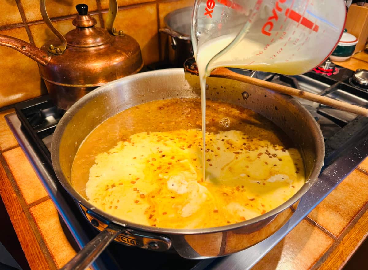 Heavy cream being poured from a pyrex measuring cup into chicken stock flecked with chopped shallot and mushroom mixture in a large metal skillet.