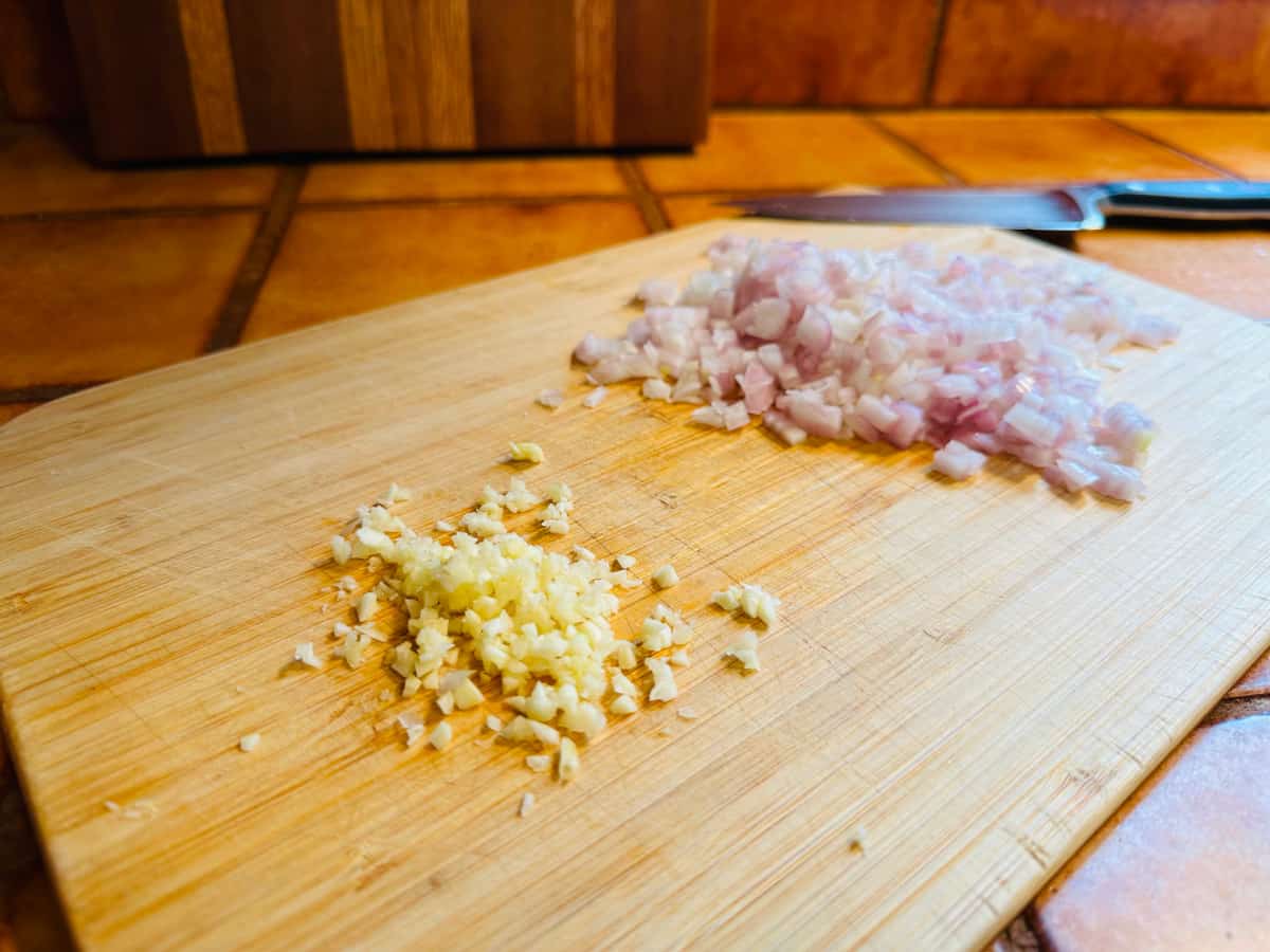 Chopped garlic and shallot on a light colored cutting board with a small chef's knife.