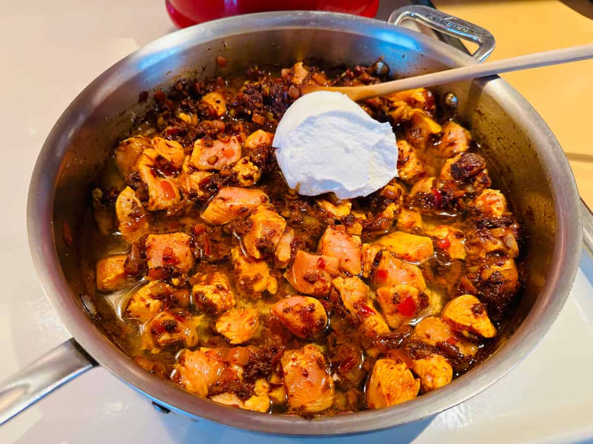 A large dollop of plain Greek yogurt on top of chicken cubes mixed with browned spices and water in a steel saute pan with a wooden spoon.