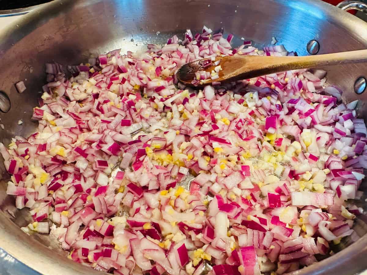 Diced red onion with minced garlic and ginger frying in a steel saute pan with a wooden spoon.