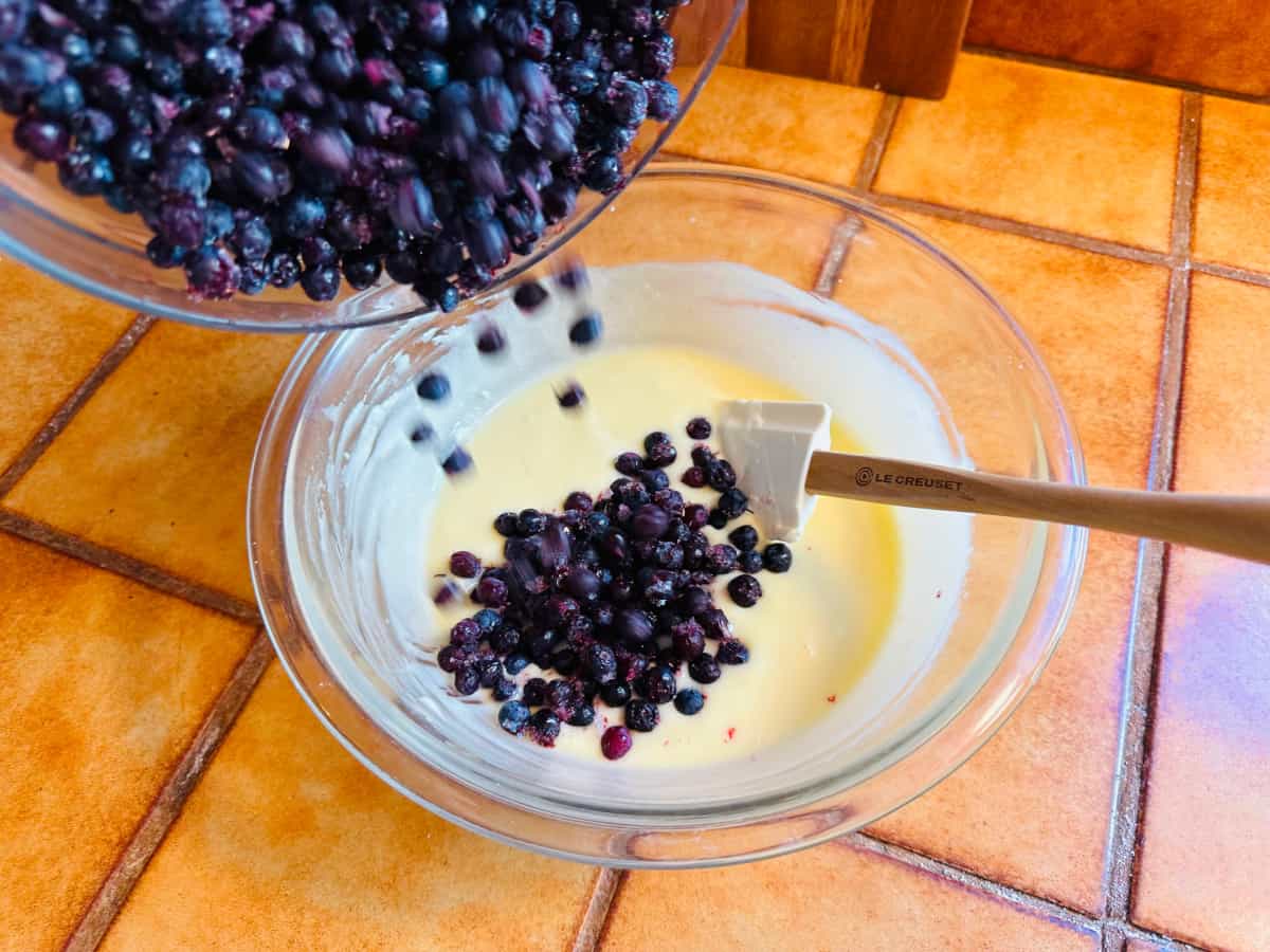 Blueberries being poured into a creamy pale yellow filling mixture in a glass bowl with a white silicone spatula.