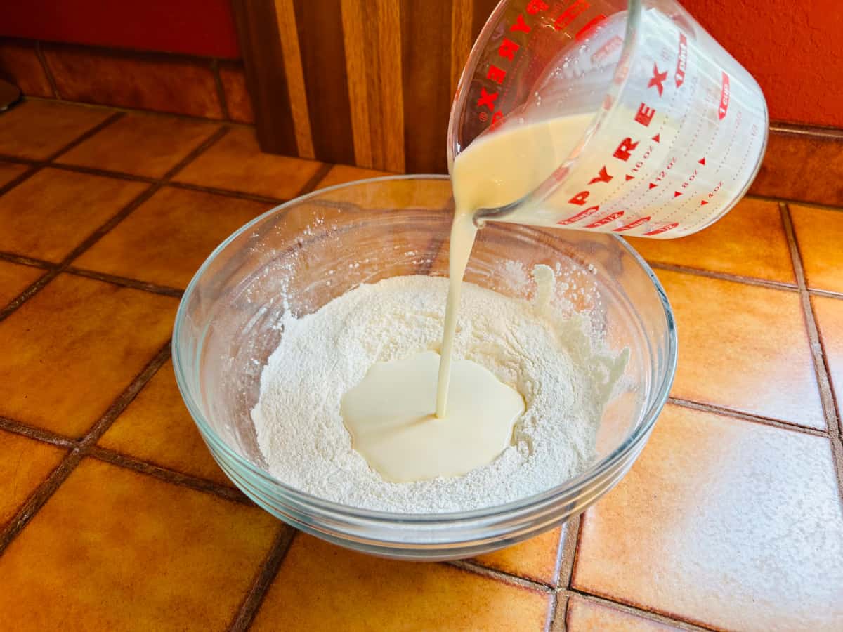 Heavy whipping cream being poured from a glass measuring cup into a glass bowl of dry ingredients.