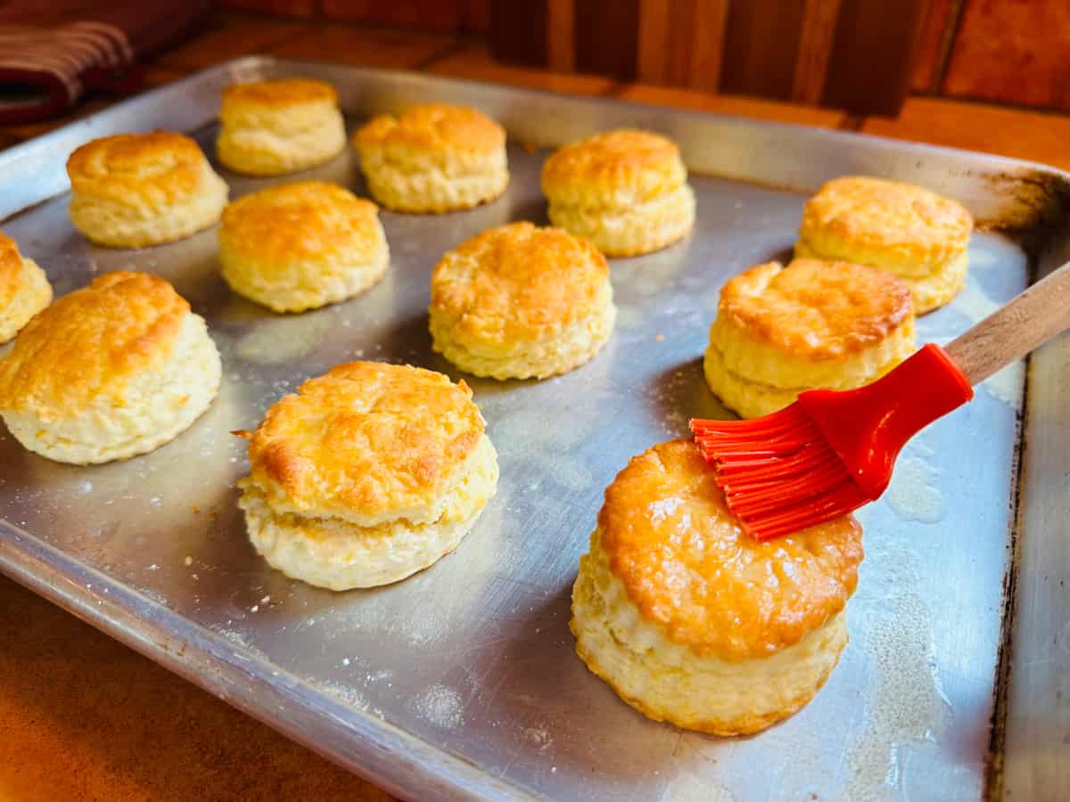 Cream biscuits on a baking sheet being brushed with melted butter by a red silicone pastry brush.