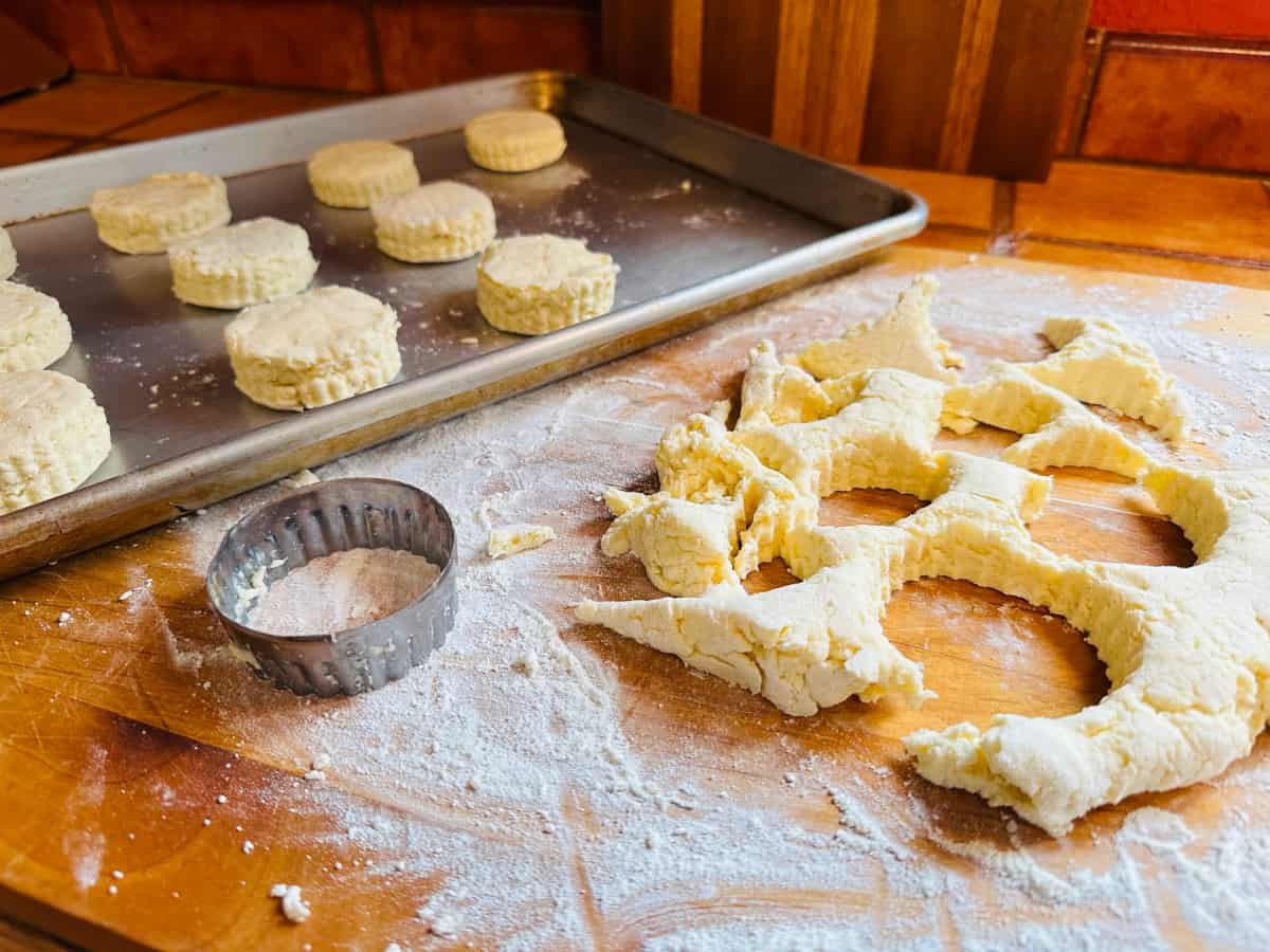Unbaked biscuits on a metal baking sheet next to a biscuit cutter and scraps on a floured wooden board.