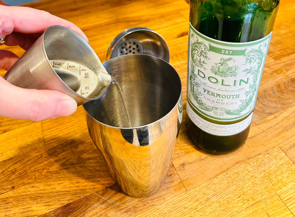 Dry vermouth being poured from a metal measuring cup into a cocktail shaker.