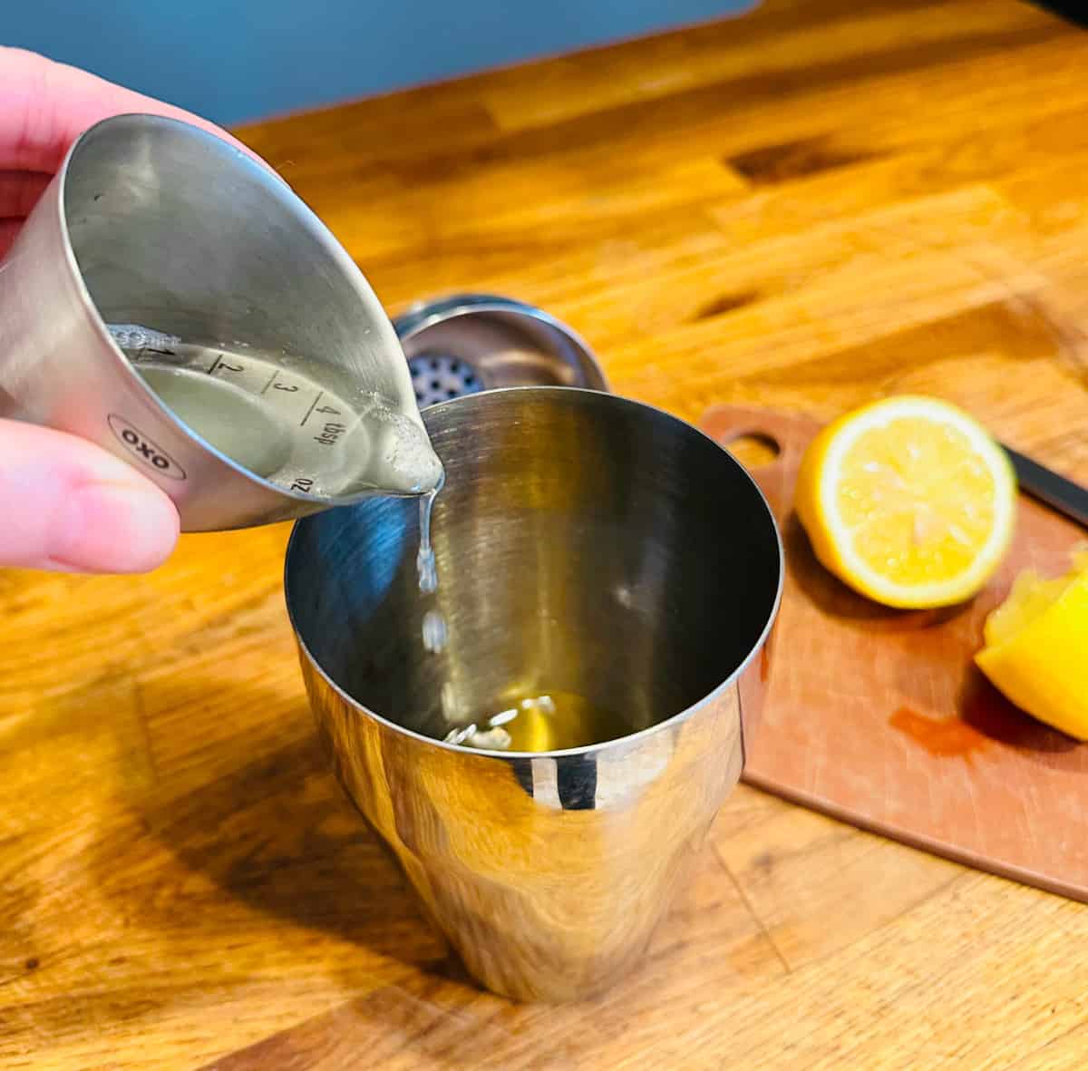 Lemon juice being poured from a metal measuring cup into a cocktail shaker.