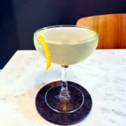 Elderflower martini with a lemon twist in a small coupe glass sitting on a dark gray coaster on a white marble table.
