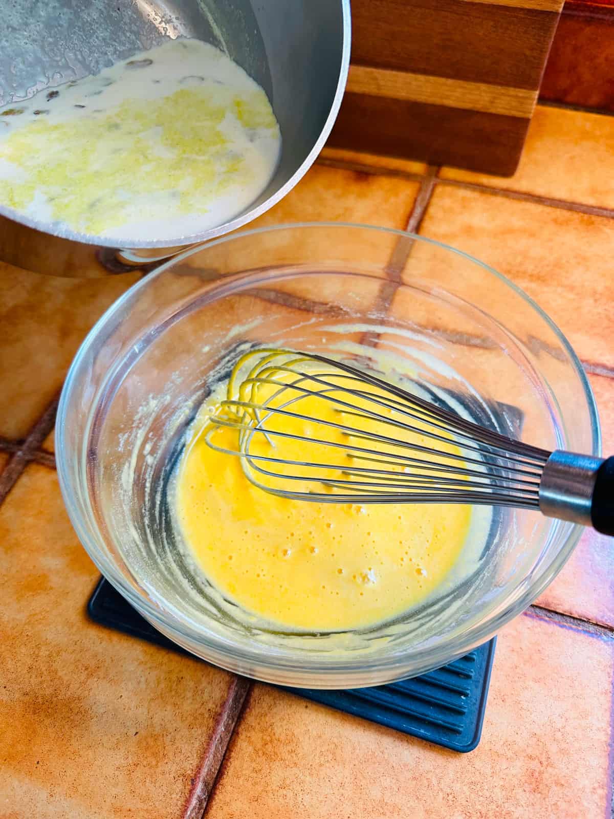 White chocolate mixture being poured from a steel saucepan into a glass bowl with yolk mixture and a whisk.