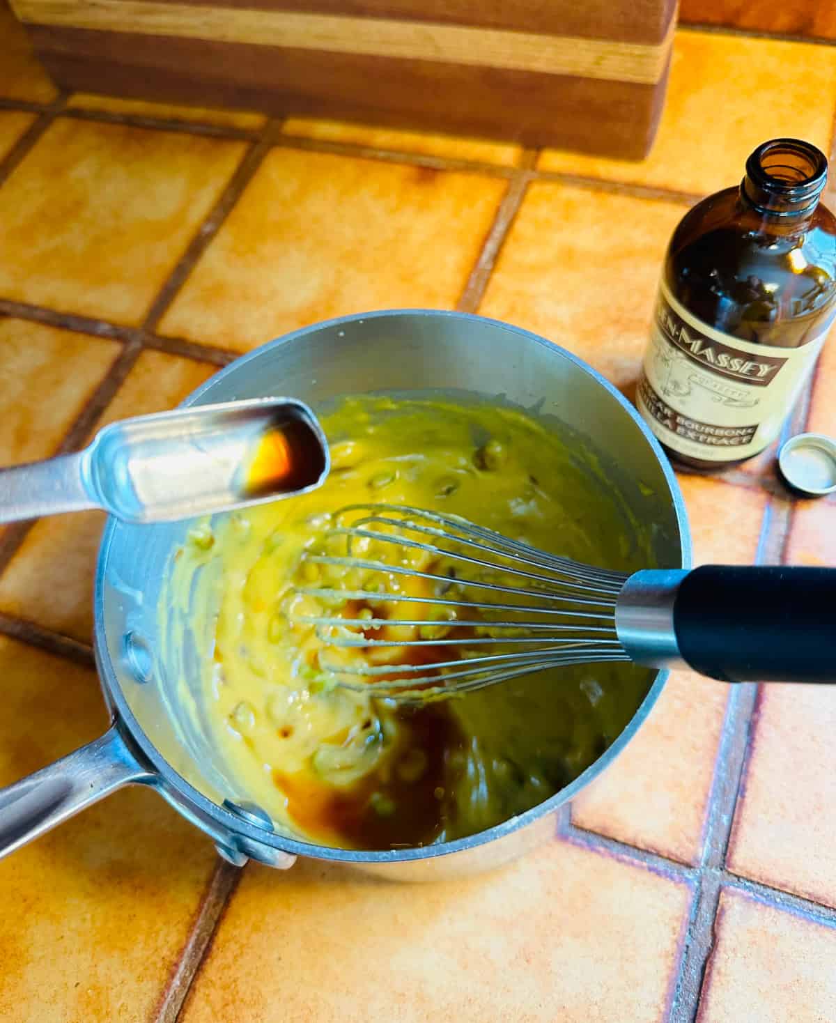 Vanilla extract being poured from a teaspoon into pistachio pastry cream in a small steel saucepan with a whisk.