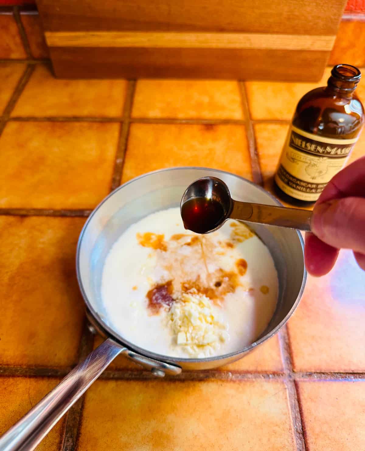 Vanilla extract being poured from a teaspoon into a small steel saucepan with half & half and white chocolate.