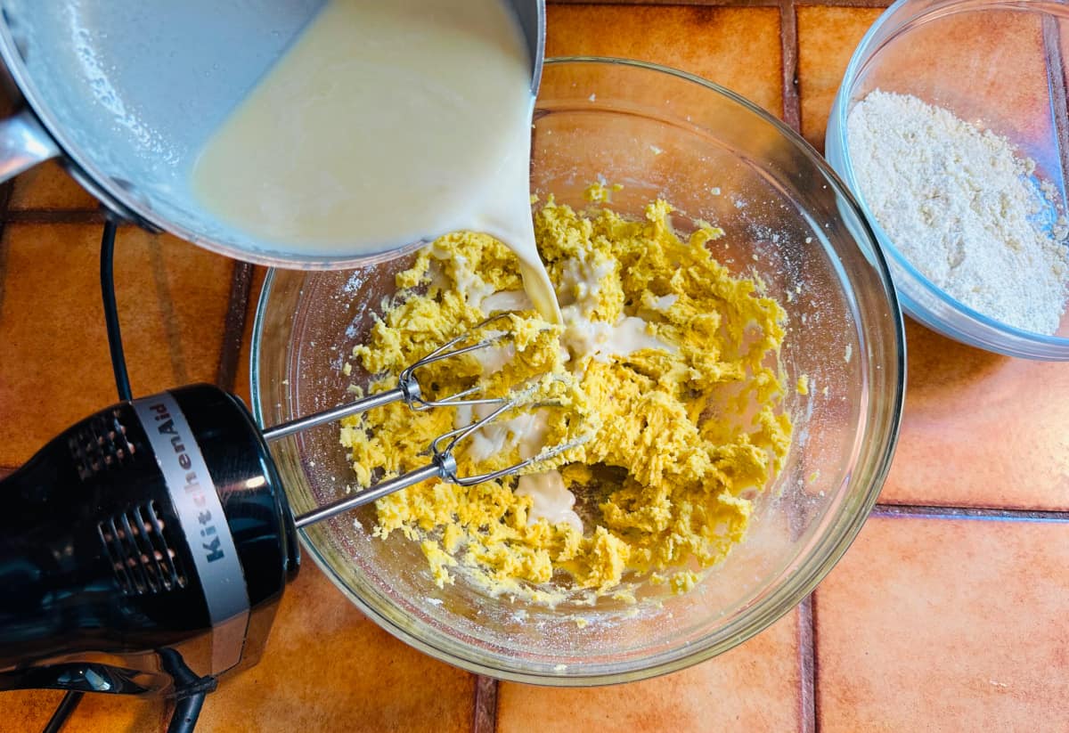 White chocolate mixture being poured from a metal saucepan into a glass bowl with cake mixture next to an electric mixer and a bowl of dry ingredients.