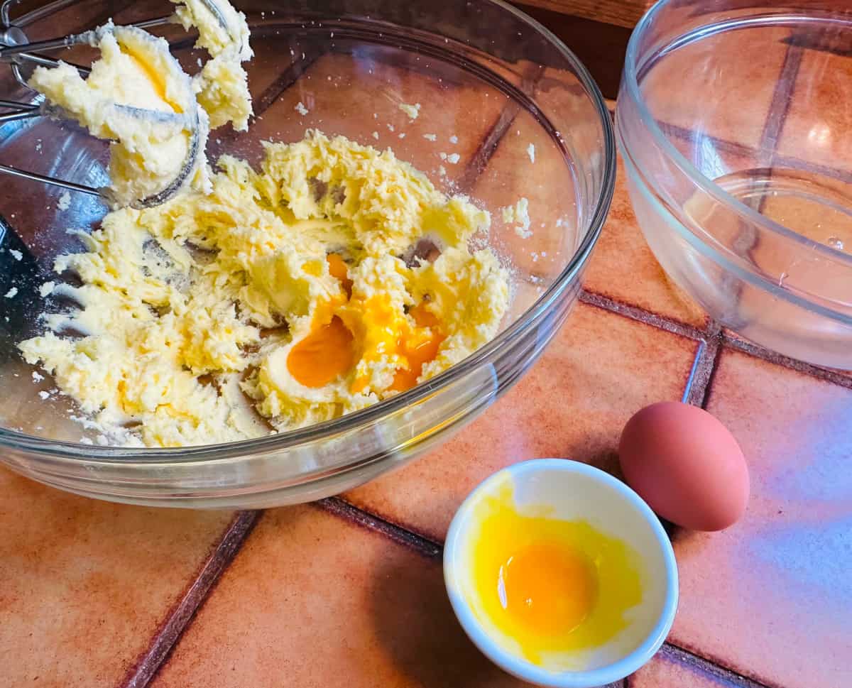 Butter, sugar, and egg yolks in a large glass bowl with beaters next to a small white bowl of egg yolk, a whole egg, and a glass bowl with egg whites.