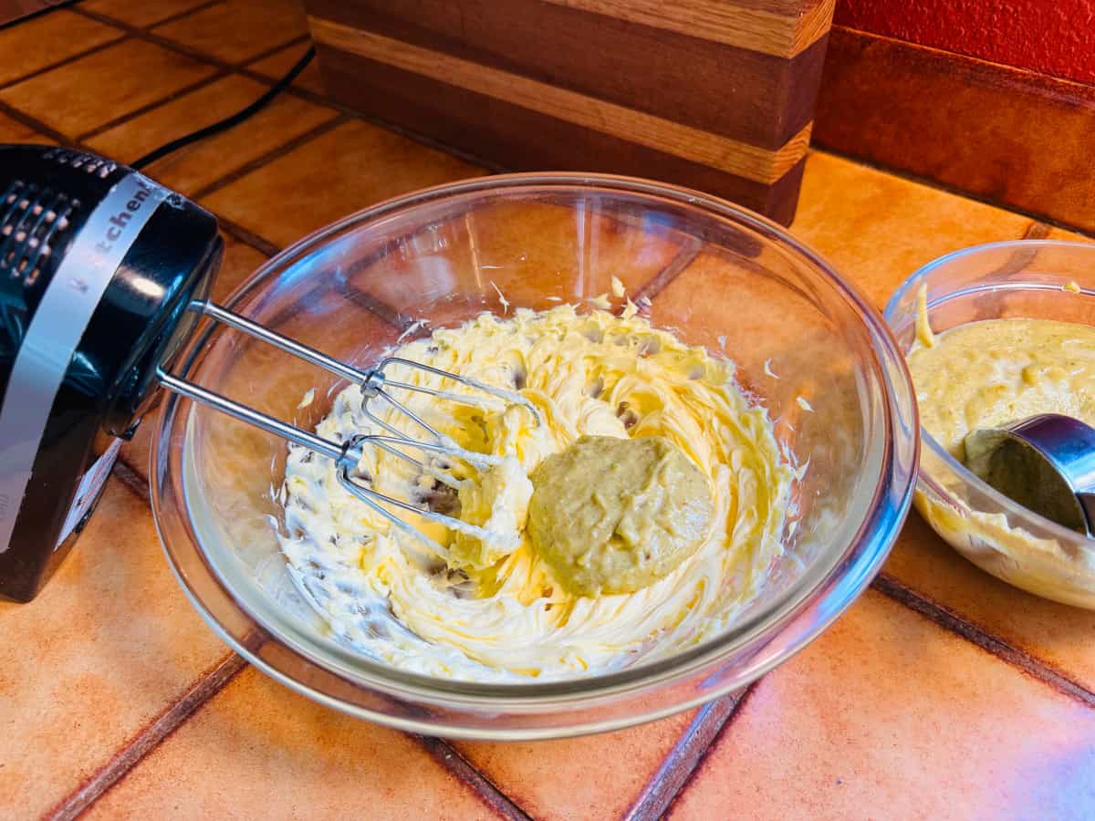 Electric mixer leaning on a glass bowl of beaten butter with a dollop of pastry cream next to a glass bowl of pastry cream with a measuring cup.