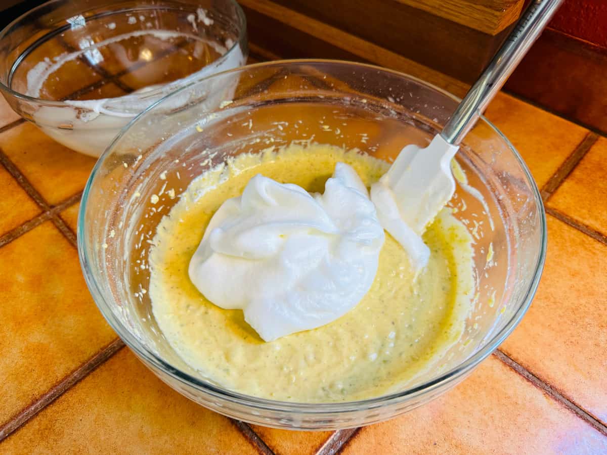 Pile of whipped egg whites on top of pale yellow batter in a glass bowl with a white silicone spatula.