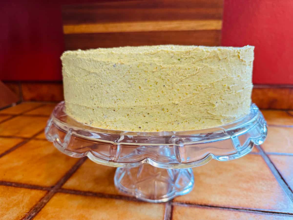 Pistachio cake frosted with buttercream on a scalloped glass cake plate.