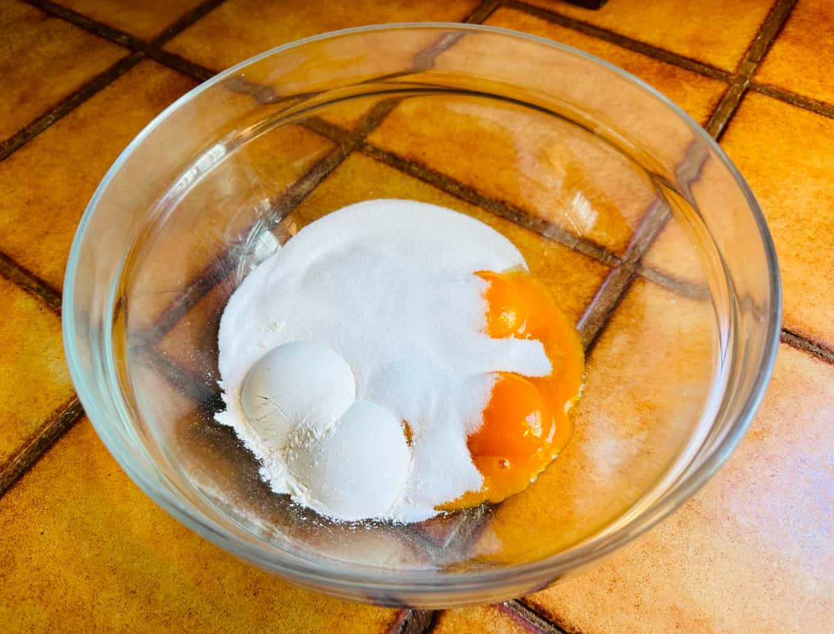 Sugar, flour, and egg yolks in a glass bowl on a terra cotta tiled counter.