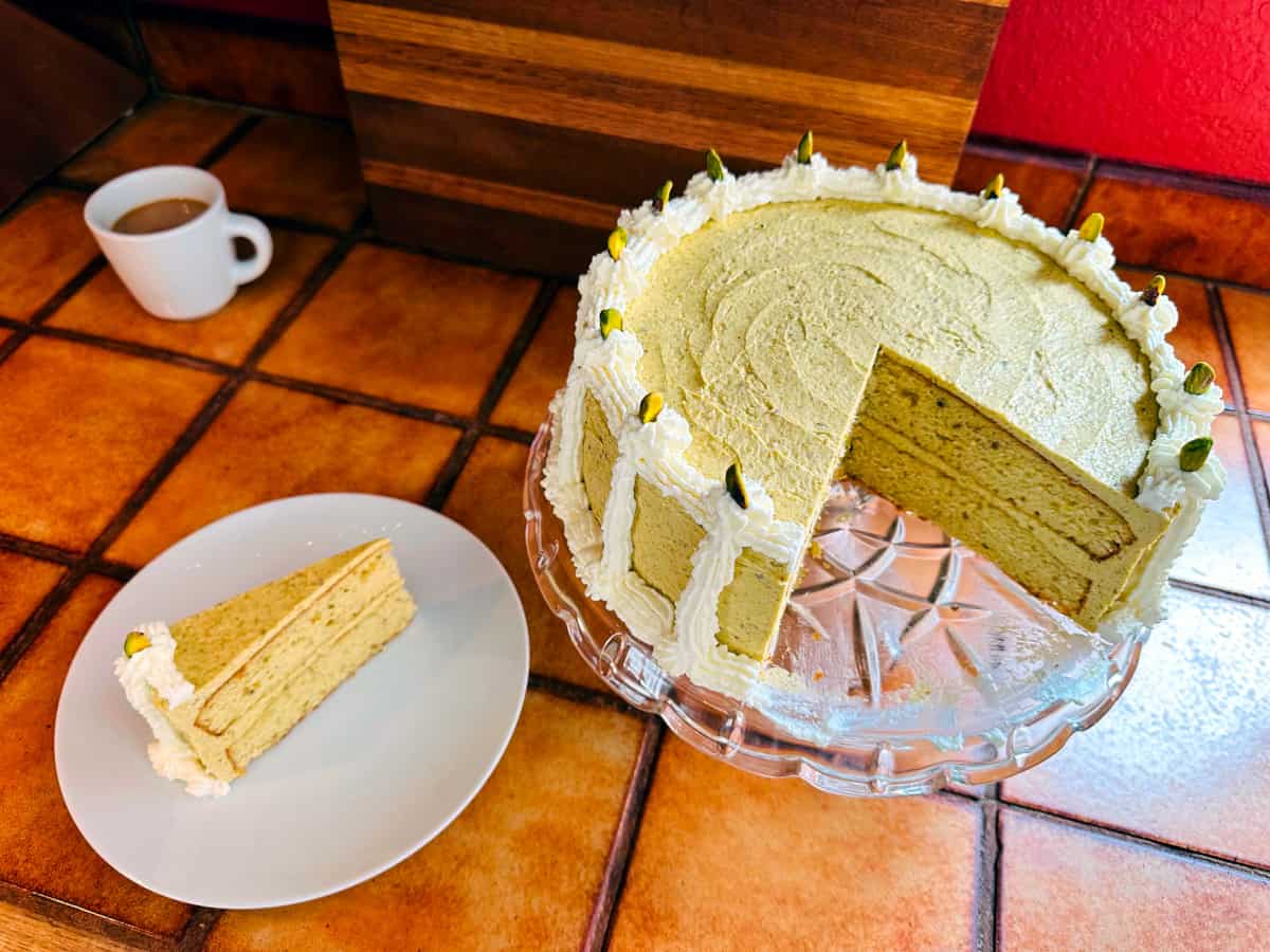 Pistachio cake with a large wedge cut out on a scalloped glass cake plate next to a slice of cake on a white plate and a white cup of coffee.