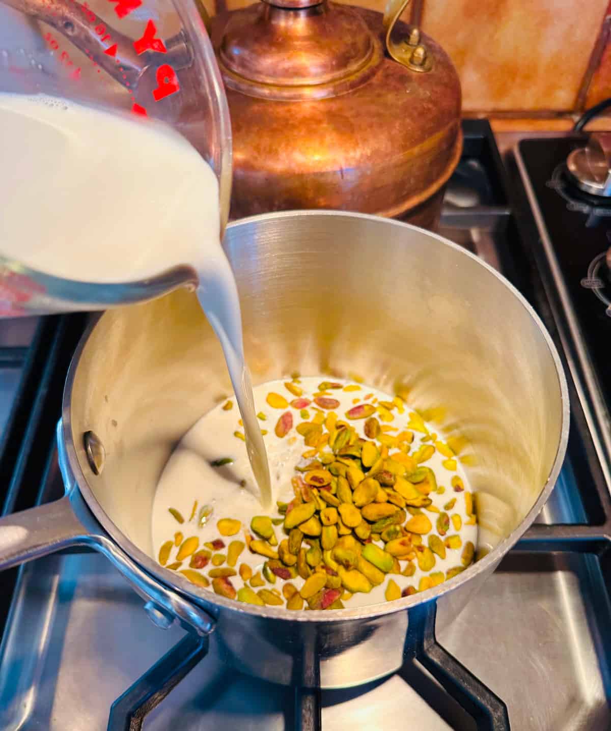 Half & half being poured from a glass measuring cup into a small steel saucepan full of pistachio nuts sitting on the stove.