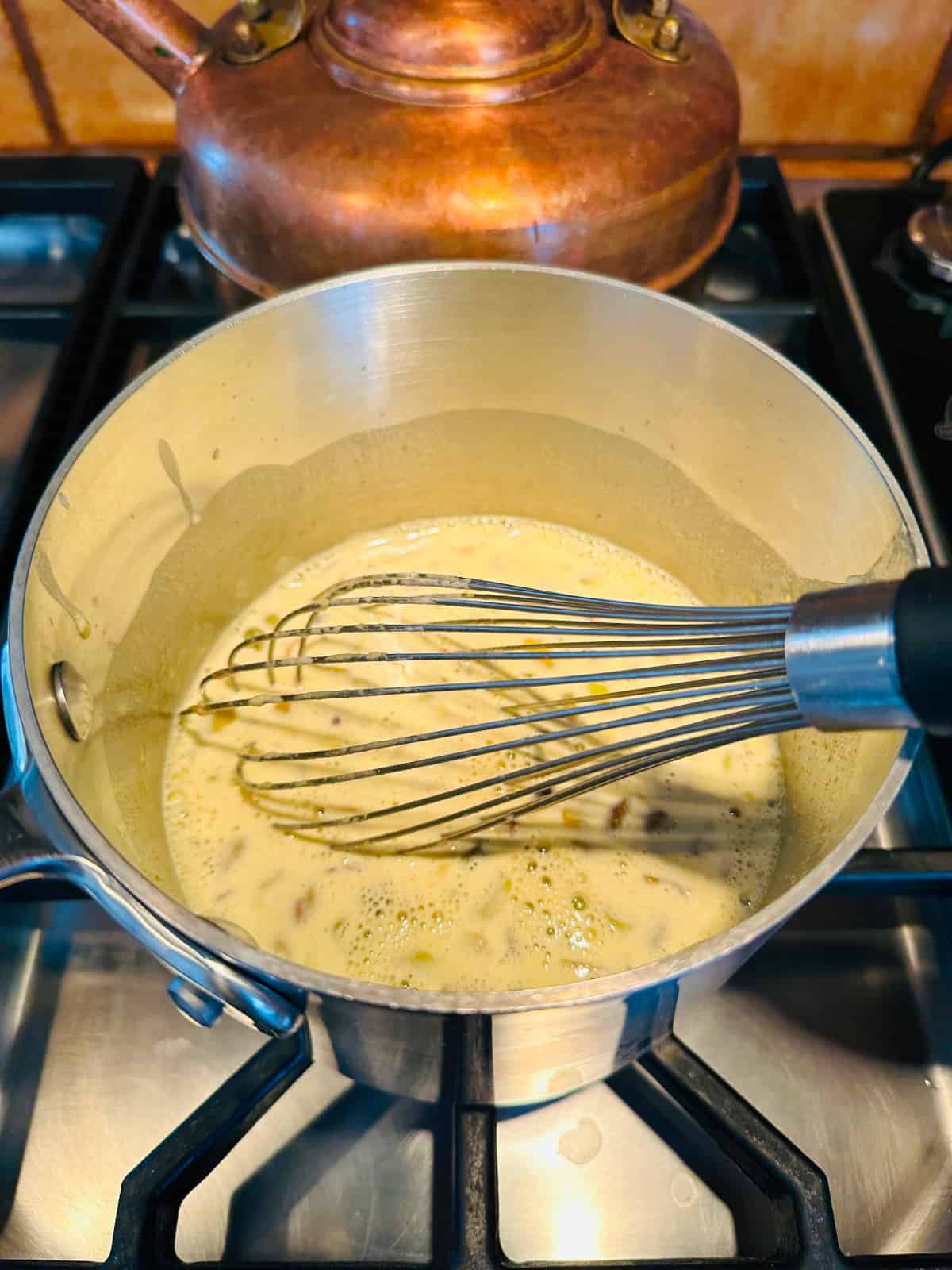 Pastry cream heating on the stove in a small steel saucepan with a whisk.