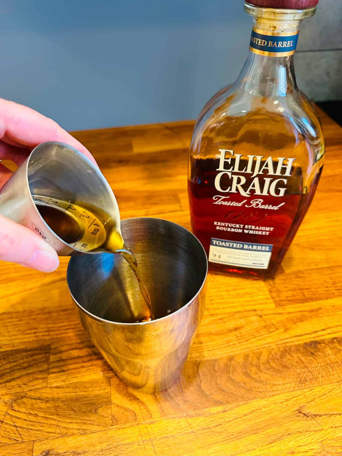 Brown liquid being poured from a steel measuring jigger into a cocktail shaker next to a bottle of Elijah Craig bourbon.