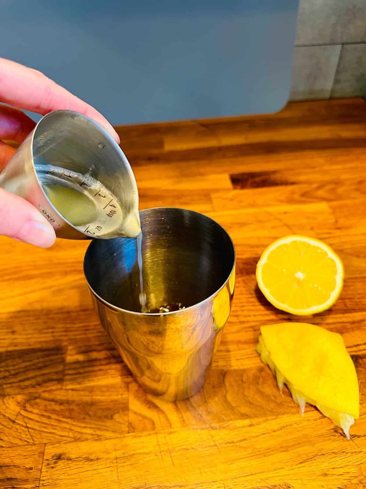 Pale yellow liquid being poured from a steel measuring jigger into a cocktail shaker next to two lemon halves.