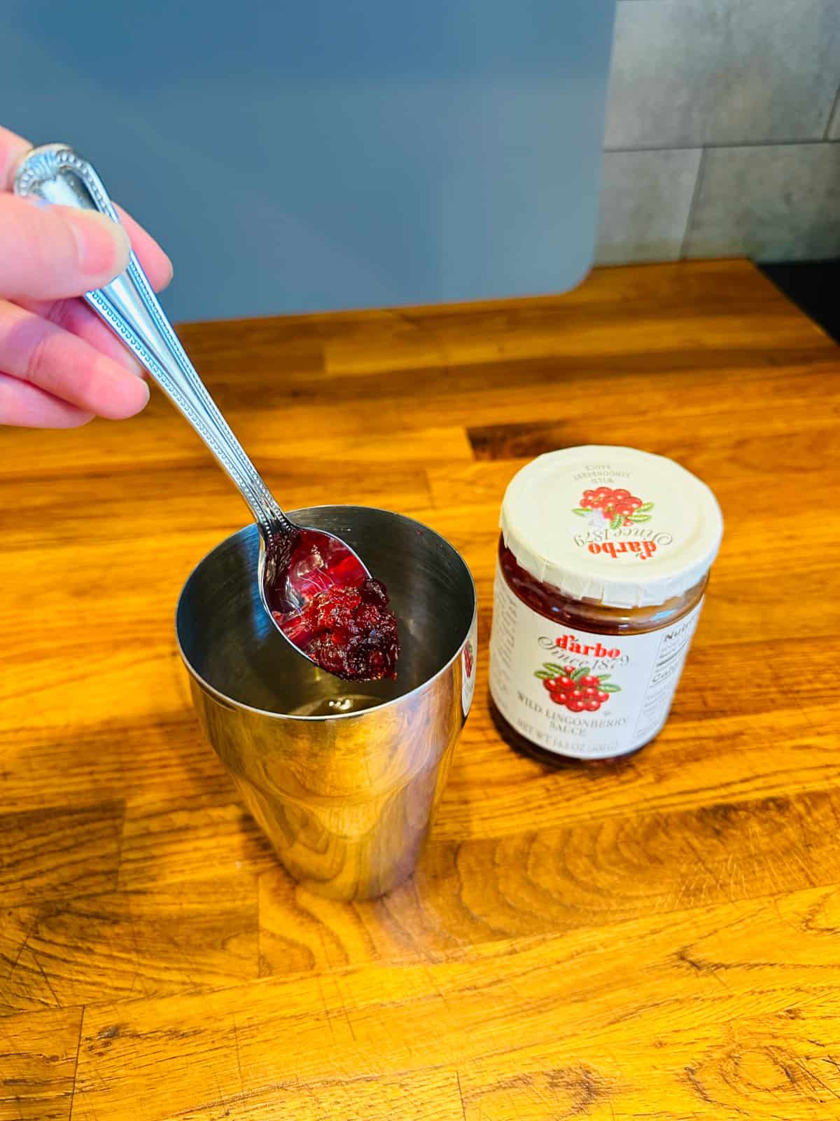 A spoonful of dark red berry sauce being added to a cocktail shaker next to a jar of wild lingonberry sauce.