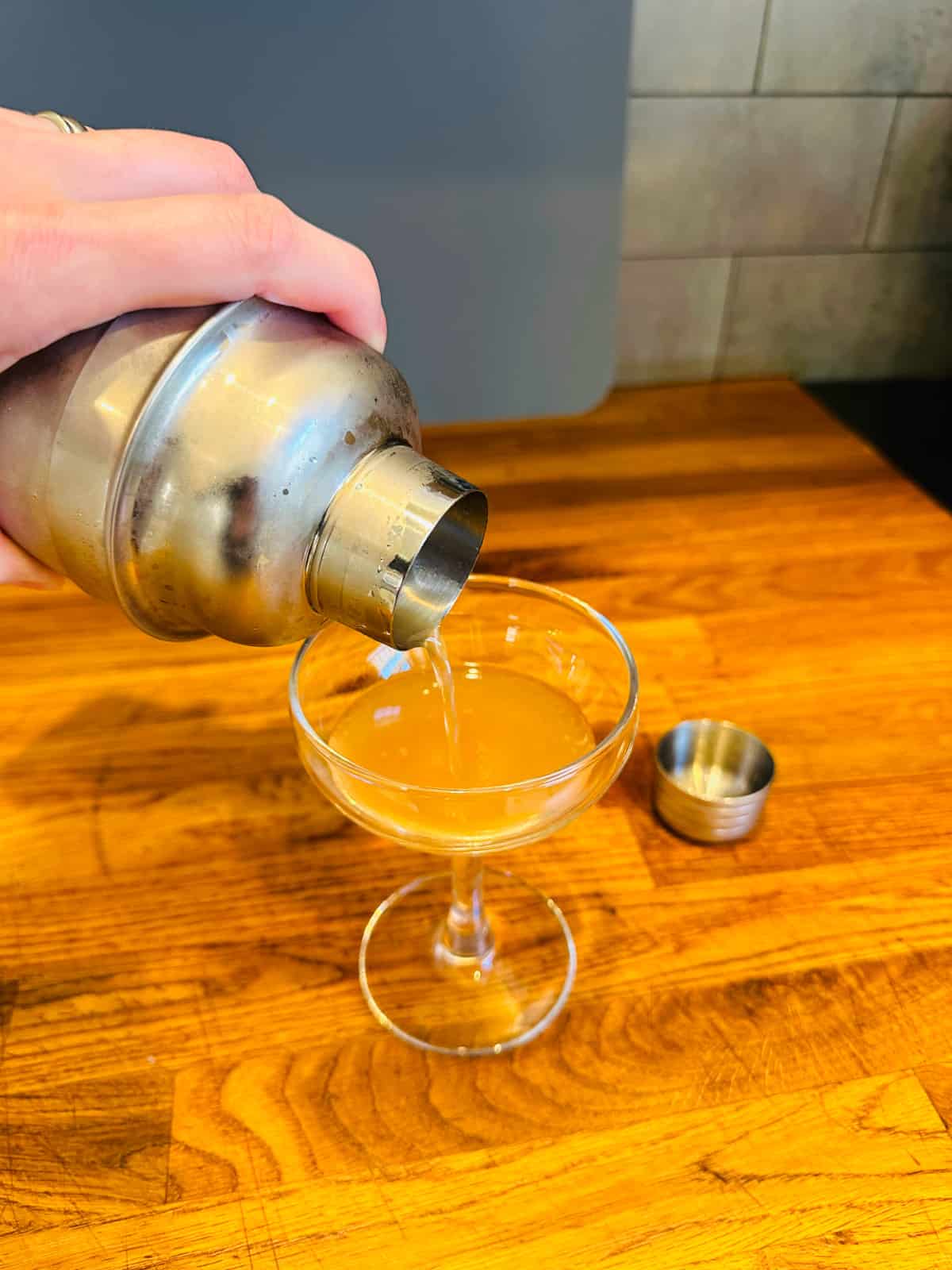 Pale orange colored liquid being poured from a cocktail shaker into a coupe glass.