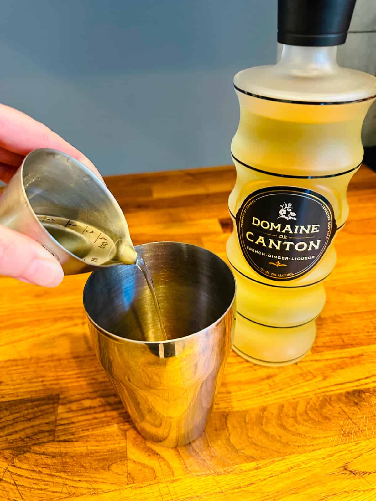 Pale yellow liquid being poured from a steel measuring jigger into a cocktail shaker next to a bottle of Domaine de Canton ginger liqueur.