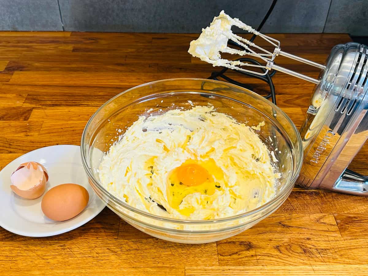 Creamed butter and sugar with one egg in a glass bowl in between an eggshell and a whole egg on a small white plate and a silver handheld electric mixer.