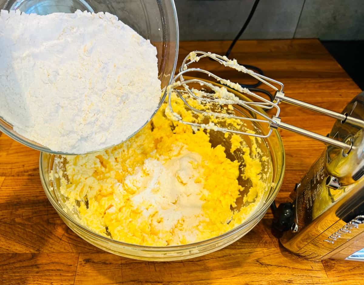 Flour being poured from a glass bowl into yellow cake batter in a glass bowl with a silver handheld electric mixer.