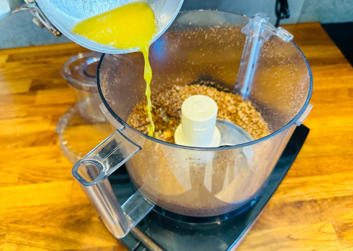 Melted butter being poured from a small saucepan into a food processor containing chopped nuts.