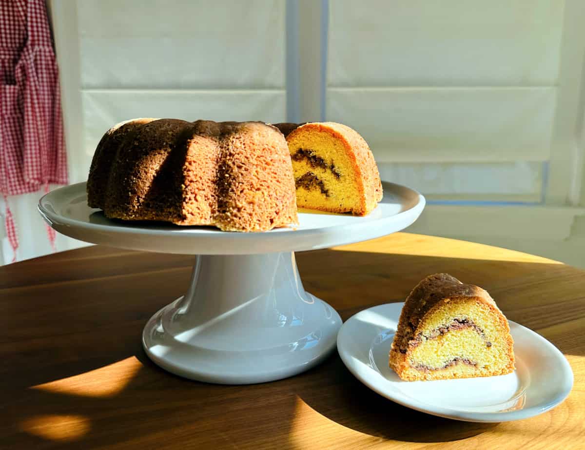 Bundt coffee cake on a white cake plate next to a slice of cake on a small white plate with a red gingham apron hanging in the corner.