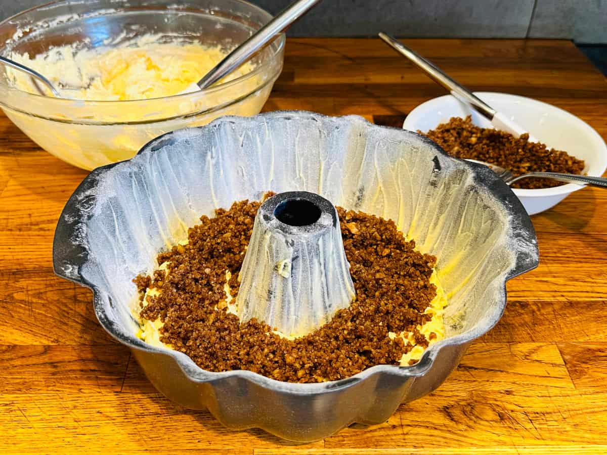 Chopped nuts sprinkled on top of a layer of cake batter in a greased and floured bundt pan in front of a bowl of batter and a bowl of chopped nuts.