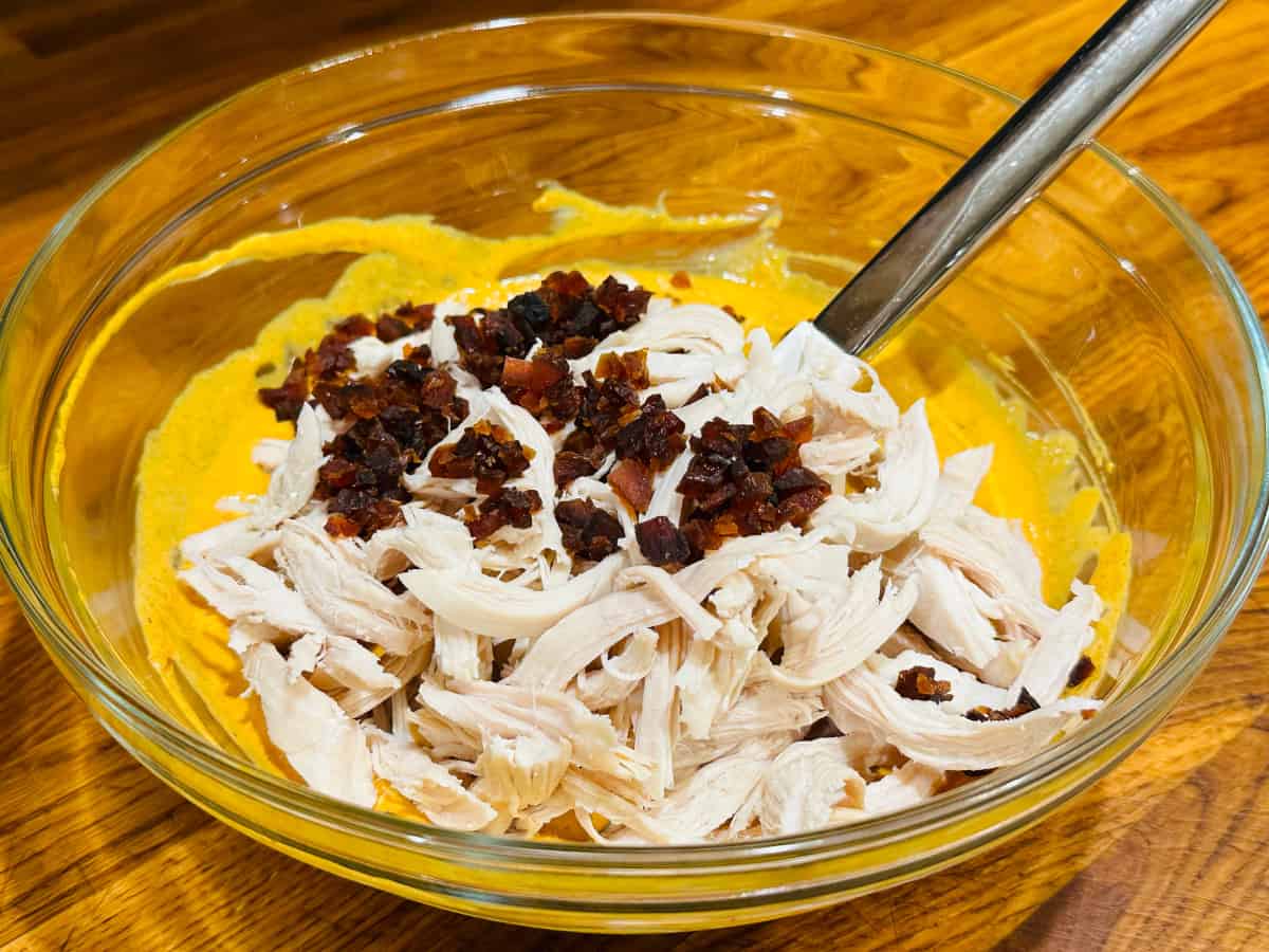 Chopped dried apricots and shredded chicken sitting on top of an orange curry mixture in a glass bowl with a steel handled spatula.