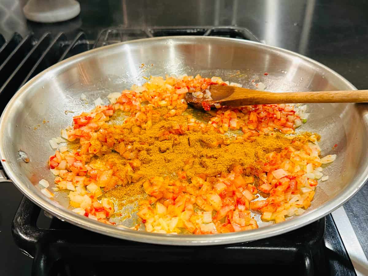Curry powder sprinkled over tomato paste coated chopped onions in a large steel saucepan with a wooden spoon.