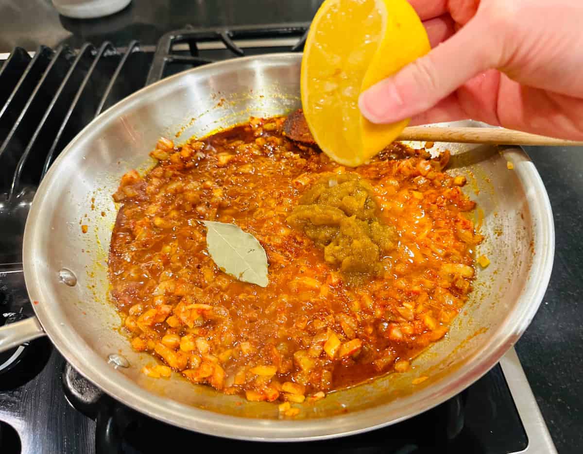 Half a lemon being squeezed over a mixture of cooked onions, red wine, mango chutney, and a bay leaf in a large steel saucepan on the stove.