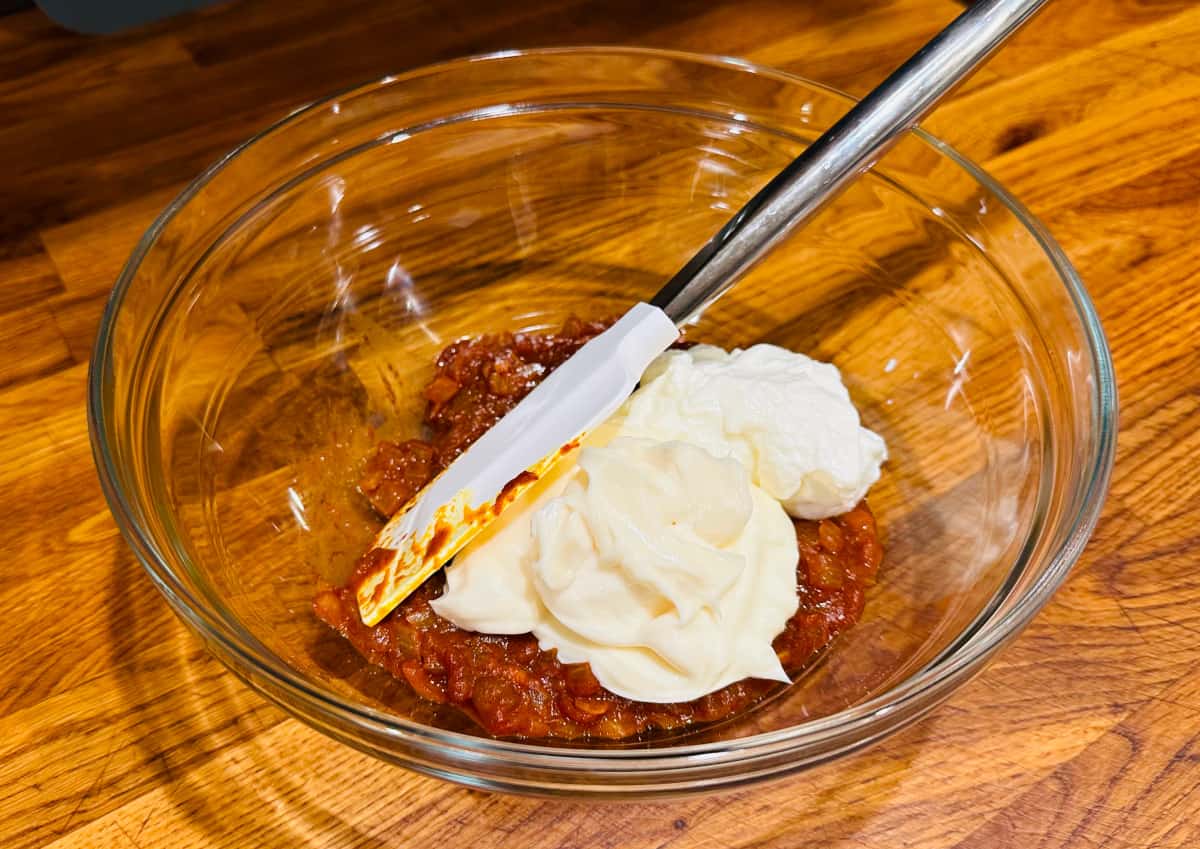 Large globs of mayonnaise and Greek yogurt on a red colored curry mixture in a glass bowl with a white silicone spatula.