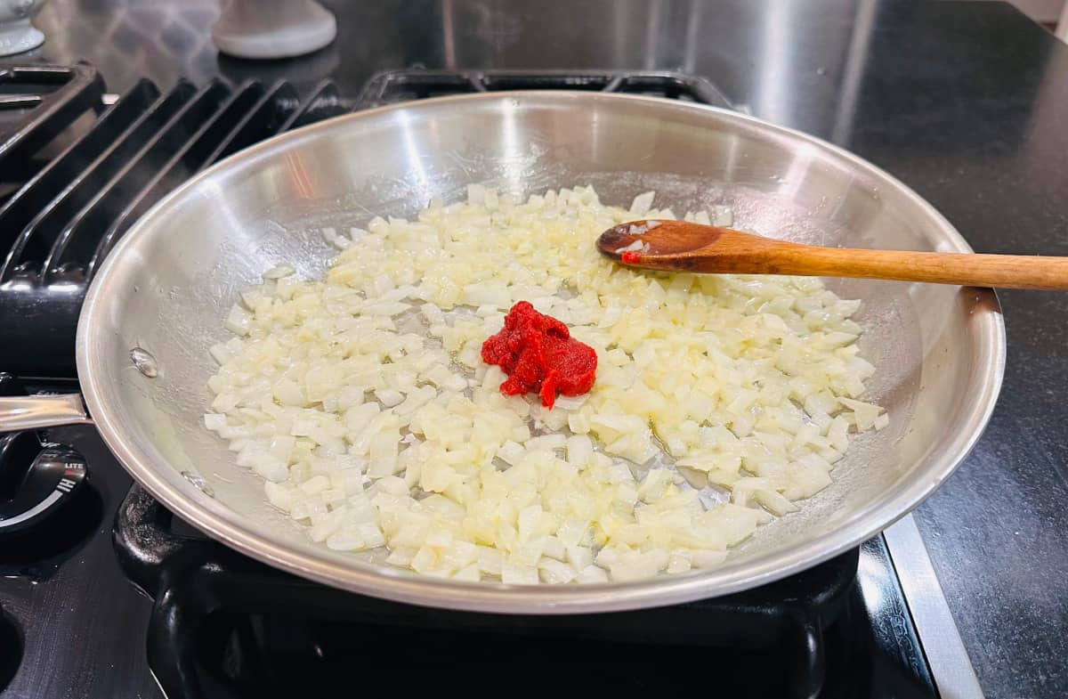 Chopped onions and a spoonful of tomato paste with a wooden spoon in a large steel saucepan on the stove.