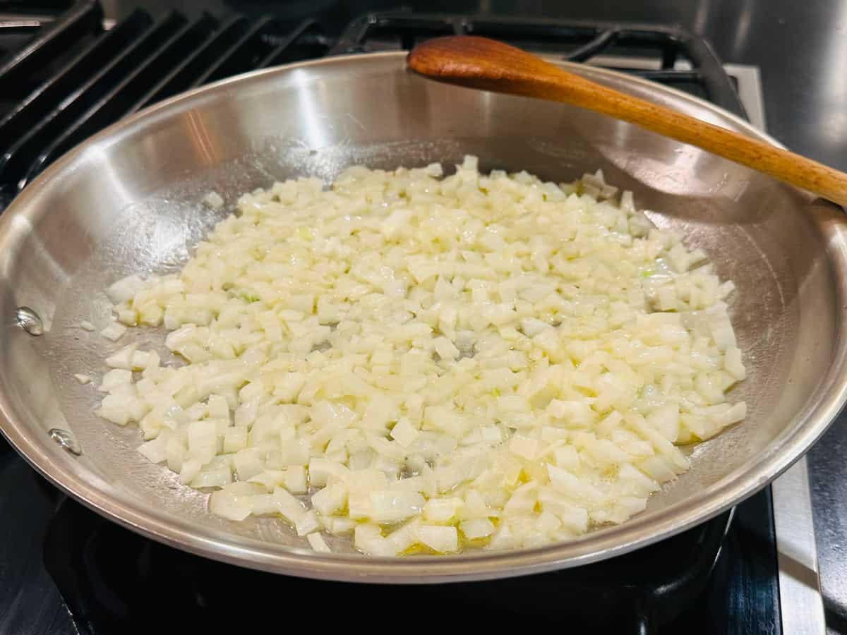 Chopped onions cooking in a large steel saucepan with a wooden spoon.