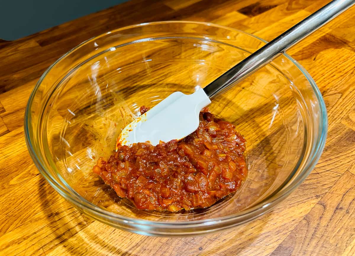 Red colored curry mixture in a glass bowl with a white silicone spatula.