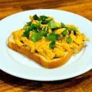 A slice of thick white sandwich bread covered with orange colored chicken salad sprinkled with bright green chopped cilantro sitting on a white plate.