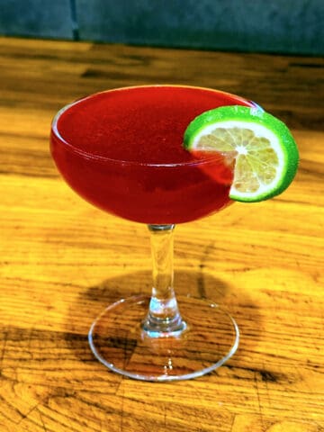 Red colored cocktail in a coupe glass garnished with a lime wheel.
